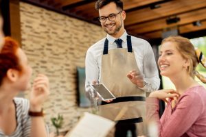 Mesero con delantal toma un pedido en una tablet mientras atiende a clientes en un restaurante